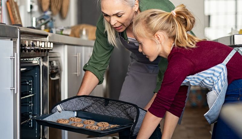 Eine ältere Frau nimmt mithilfe eines Hitzeschutzes ein heißes Blech mit gebackenen Keksen aus einem Ofen. Ein Mädchen hält die Ofentür offen - ihre nackten Unterarme kommen dabei dem Blech gefährlich nahe.