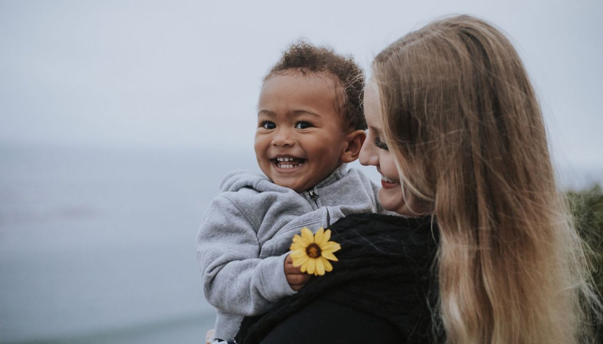 Junge blonde Frau mit dunkelhäutigem Baby auf dem Arm.