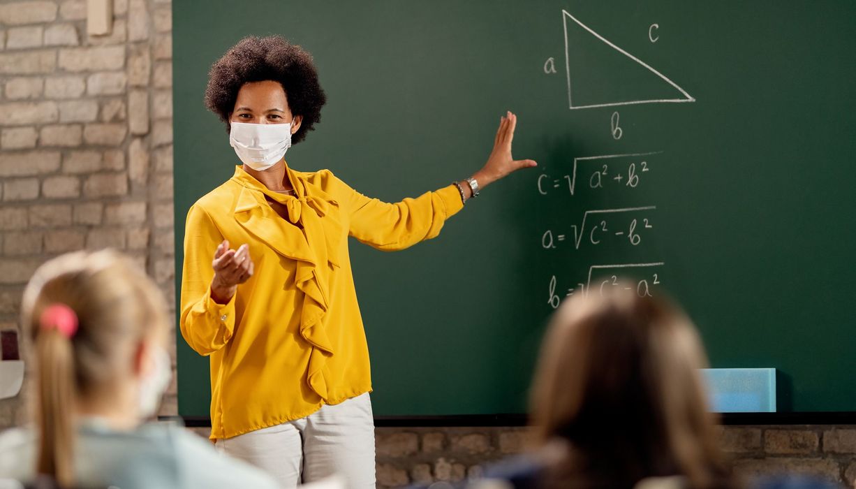 Lehrerin mit Maske steht an der Tafel und erklärt ihren Schülern eine Matheaufgabe.