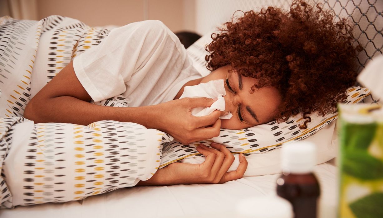 Junge Frau mit dunklen Locken, liegt im Bett und putzt sich die Nase.