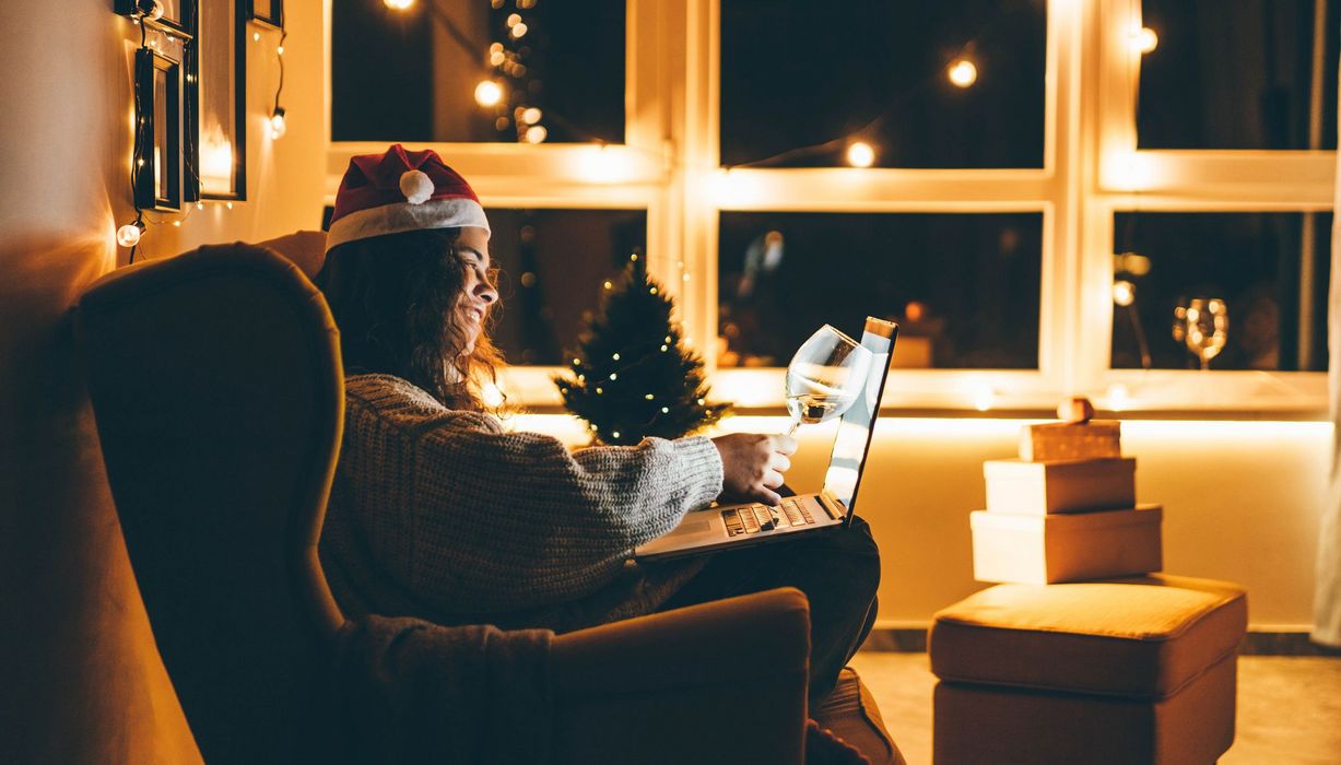 Junge Frau, sitzt auf der Couch, Laptop auf dem Schoß, lächelt in die Kamera und stößt mit einem Glas Wein an.