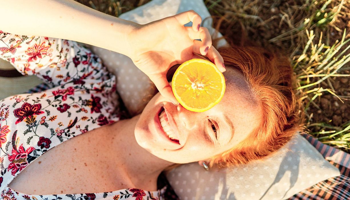 Junge Frau, liegt auf einer Wiese und hält seine Orange vor ein Auge.