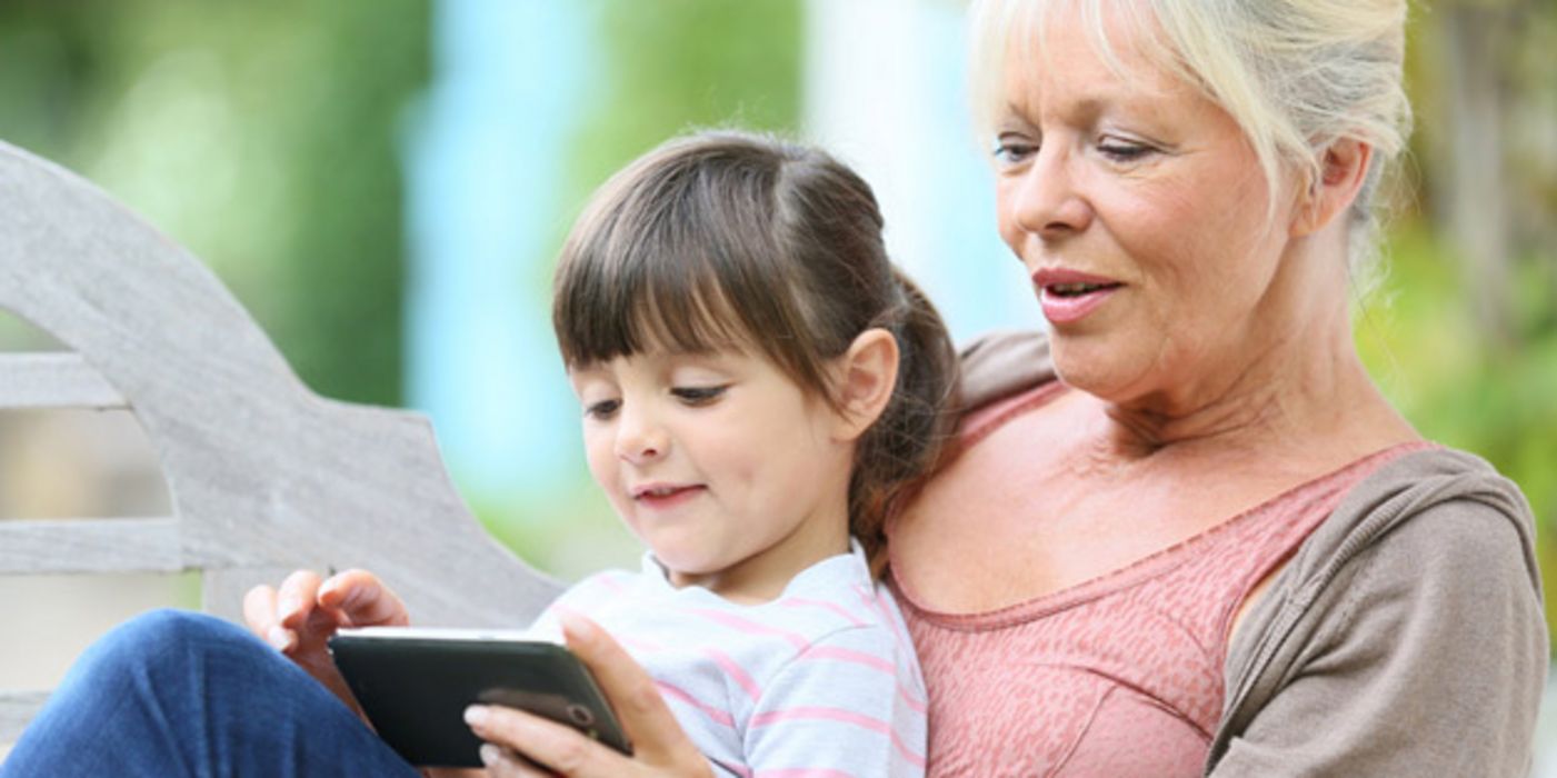 Im Freien: Attraktive Frau auf einem Gartenstuhl/Liegestuhl, um die 60, graues, zurückgebundenes Haar, auf dem Schoß ein Mädchen, ca. 6 Jahre alt. Frau zeigt ihr etwas auf dem Smartphone