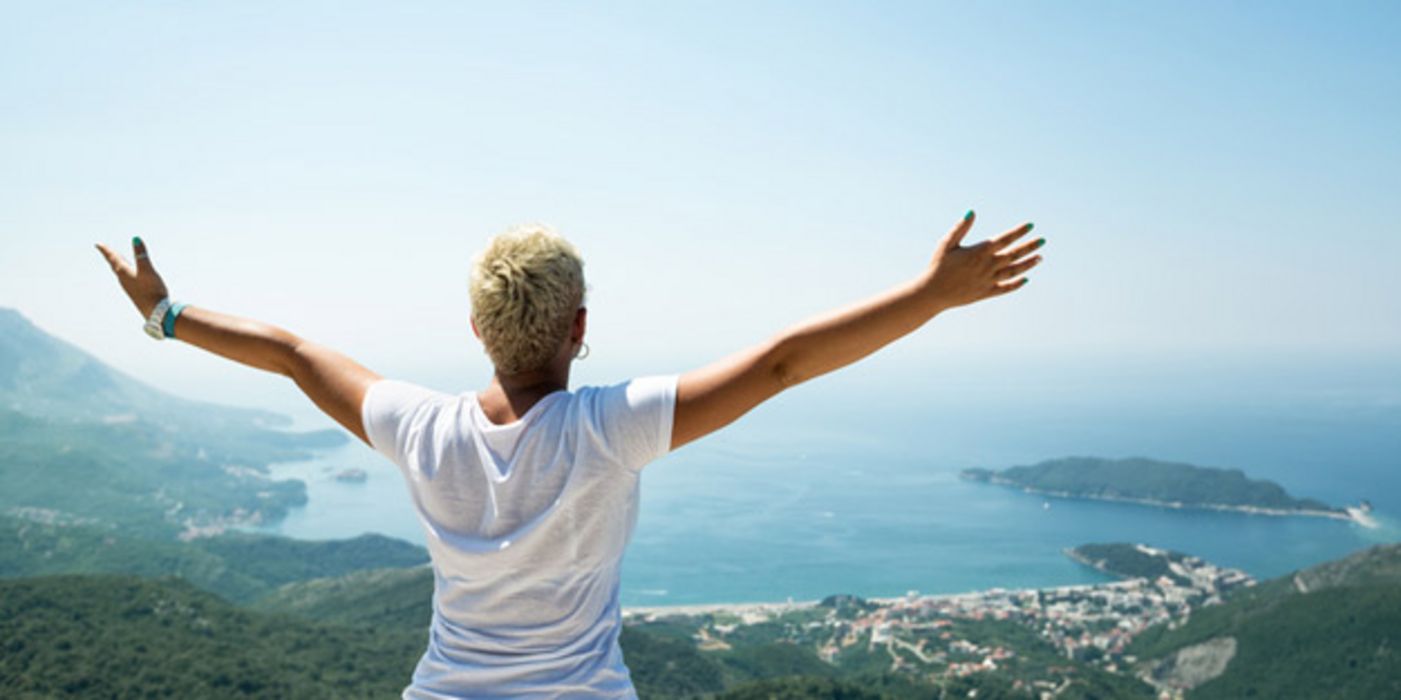 Rückenansicht: Frau, kurze, blonde Haare, weiße Bluse, auf Berg stehend auf Meer und Inseln schauend, ausgebreitete Arme