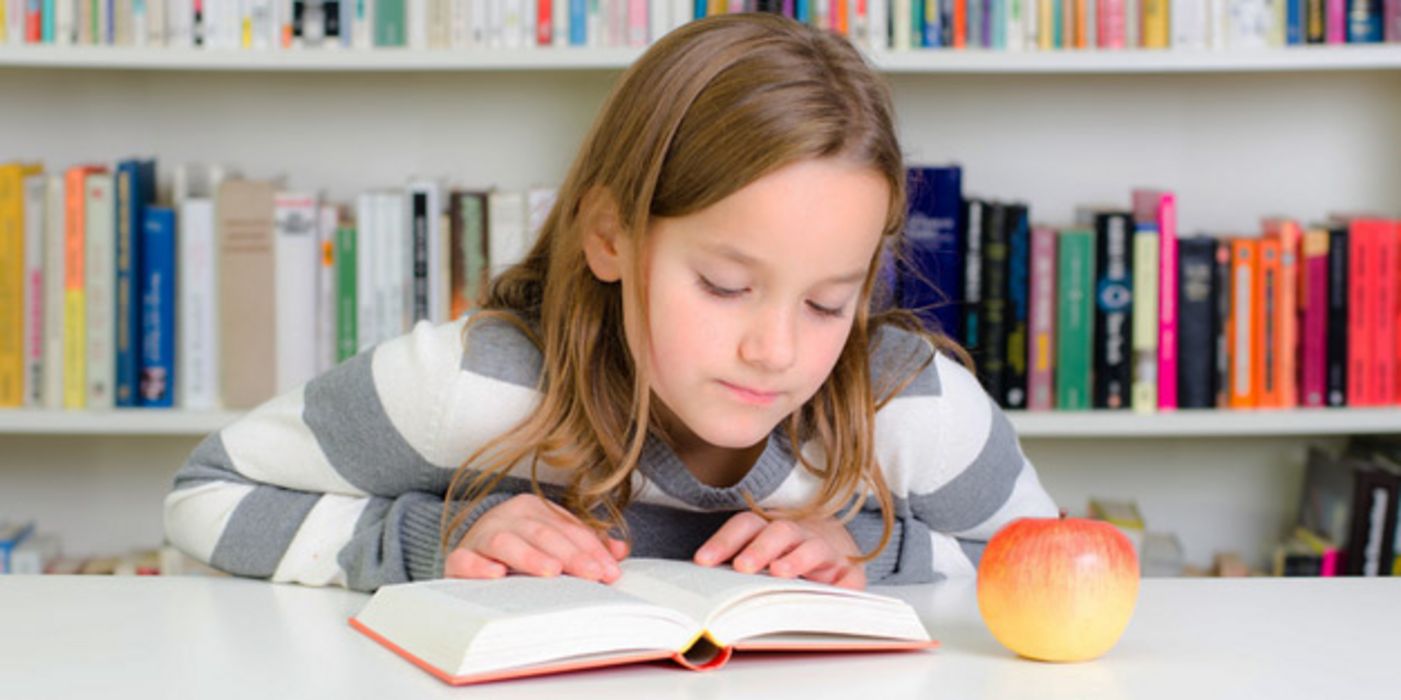 Ca. 12-jähriges Mädchen beim Lesen eines Buches, das vor ihr auf dem Tisch liegt. Hinter sich eine Bücherwand