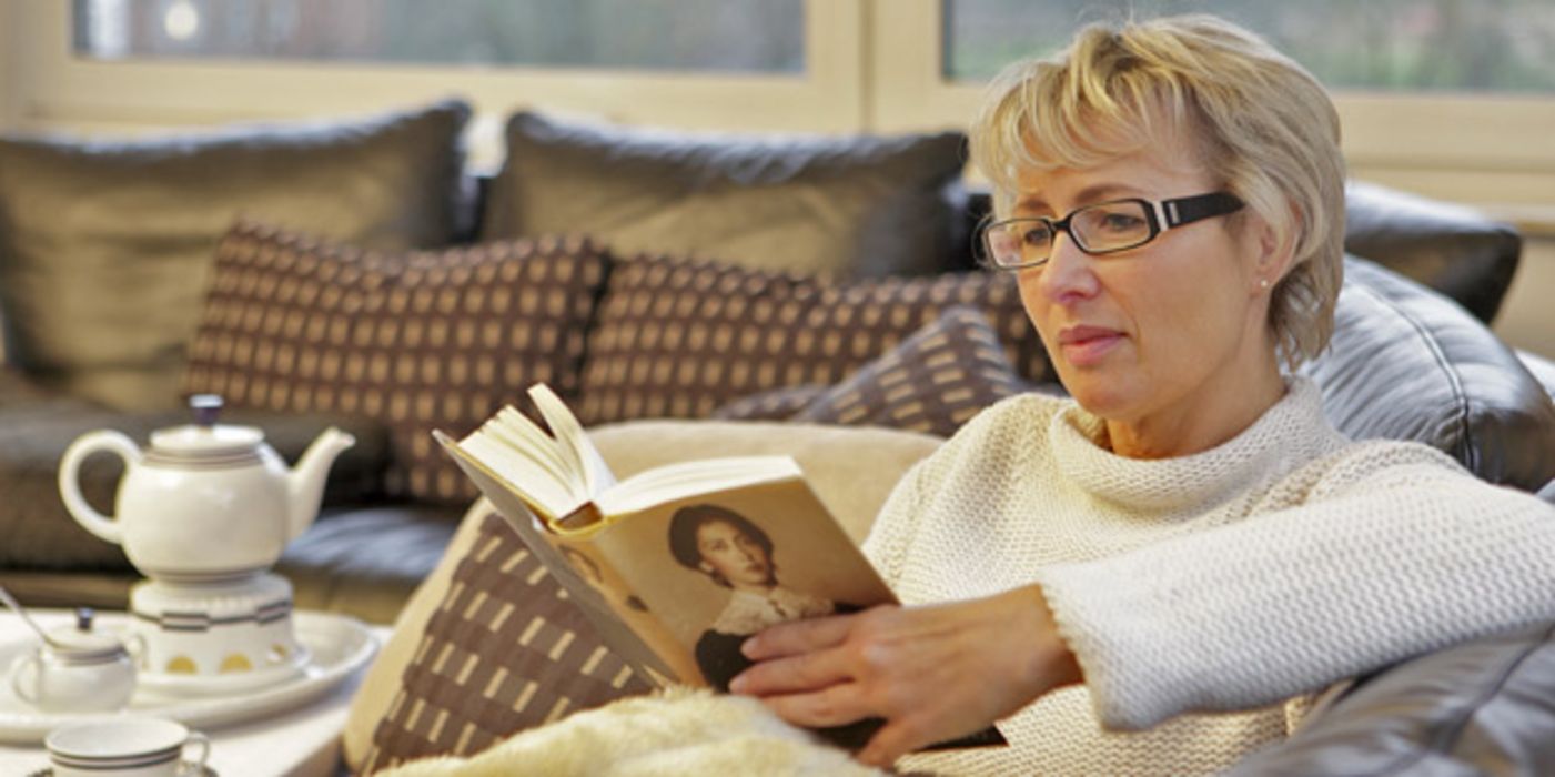 Frau in den mittleren Jahren mit Kurzhaarschnitt und Brille sitzt auf einer Couch und liest ein Buch