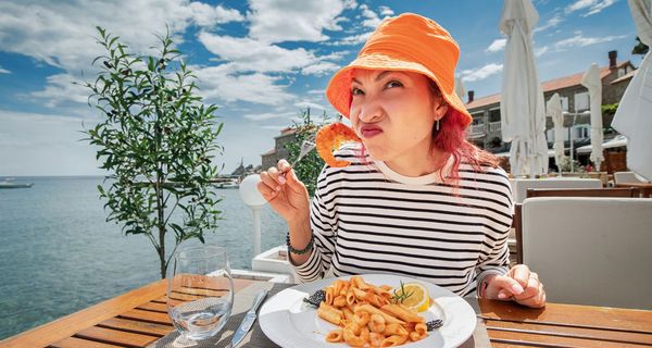 Frau mit orangem Hut isst Garnelen und rümpft skeptisch die Nase.