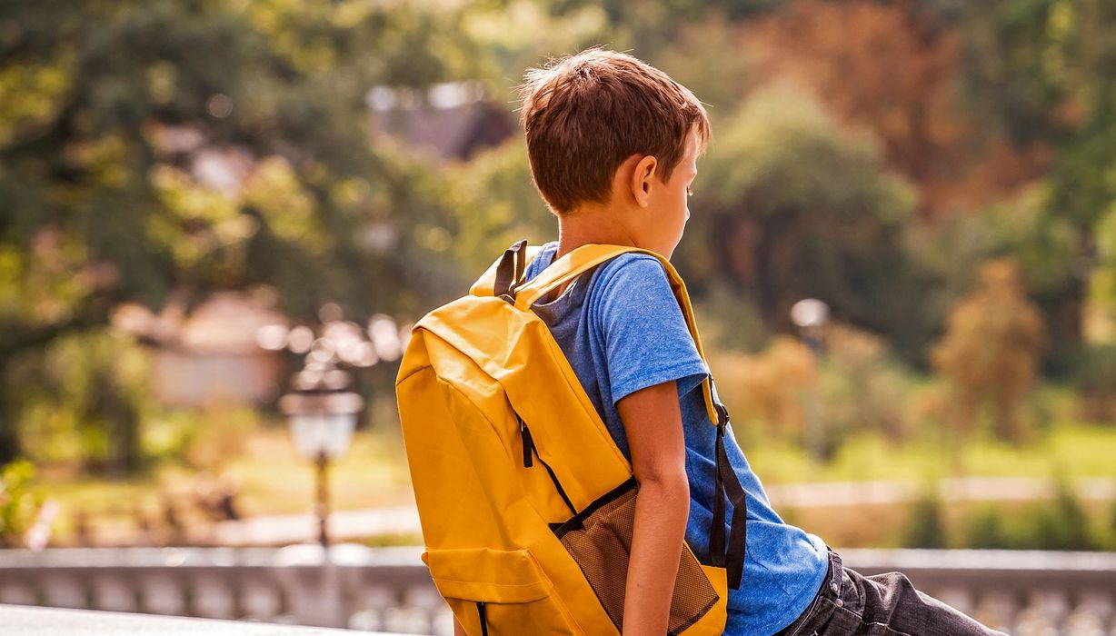 Junge mit Schulranzen, sitzt allein auf einer Mauer, Rückenansicht.