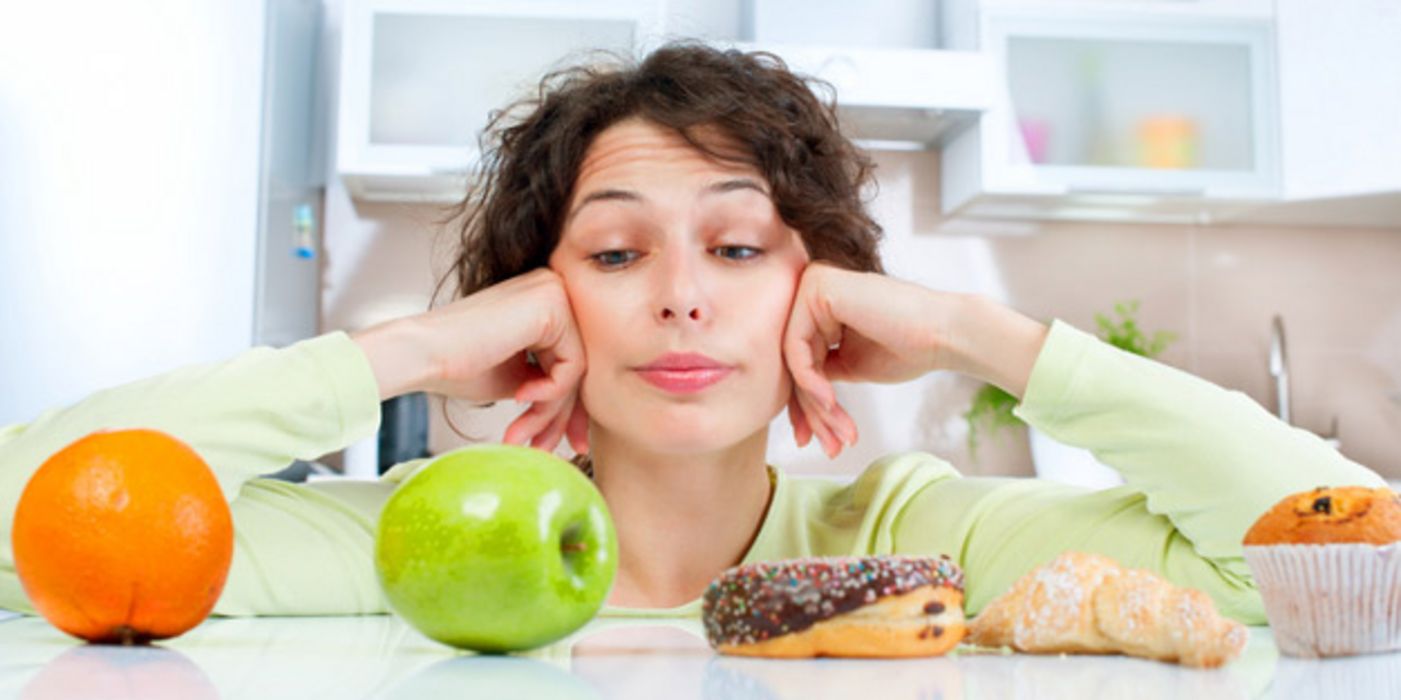 Junge dunkelhaarige Frau, Ellenbogen auf Tisch, Kopf auf Hände gestützt, schaut zweifelnd über Tischkante auf aufgereihtes Obst und Donut, Croissant und Muffin