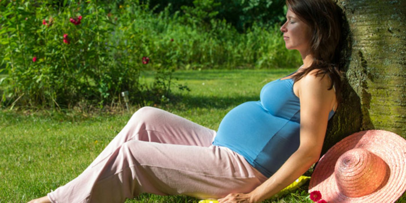 Sommerfoto: Schwangere Frau, blaues Shirt, rosa Freizeithose, Strohhut neben sich, lehnt an einem Baum auf einer Wiese und genießt die Sonne