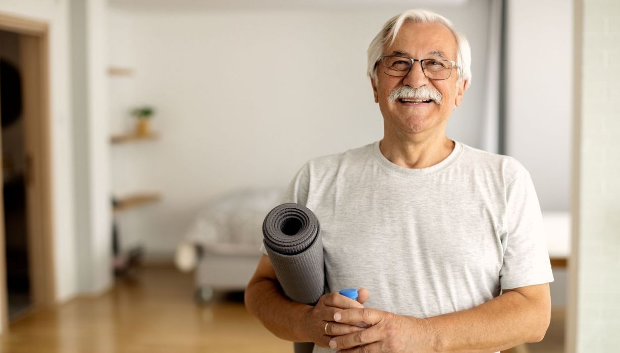 Senior, zu Hause mit Yogamatte, lächelt in die Kamera.