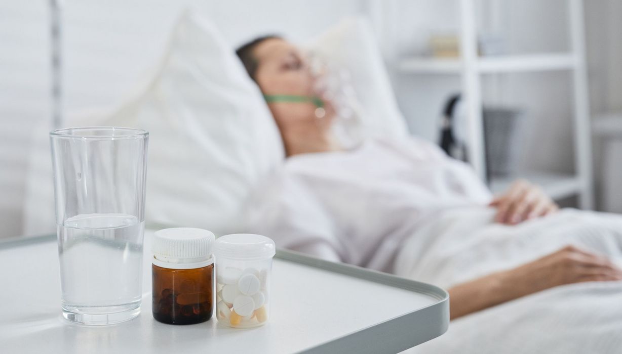 Frau im Krankenhausbett, im Vordergrund Tabletten und Wasser.