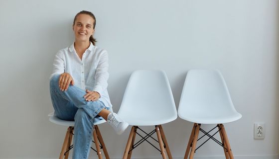 Eine junge Frau in weißem Hemd und Jeans sitzt auf einem Stuhl im Wartebereich. Sie lächelt.