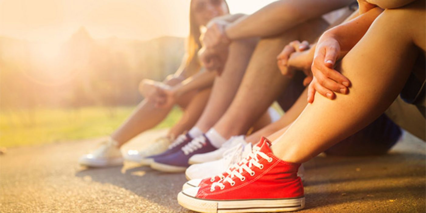 Nackte Beine von jungen Frauen die am Boden sitzen in Abendlicht, alle mit Sneakers (Chucks?), im Vordergrund rote mit weißen Sohlen