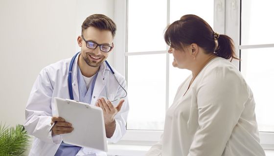 Ein Arzt zeigt lächelnd einer Frau mit Übergewicht etwas auf einem Klemmbrett.