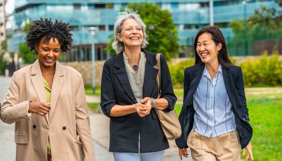 Drei Frauen in geschäftlicher Kleidung gehen spazieren, im Hintergrund eine Grünfläche und ein Bürogebäude.