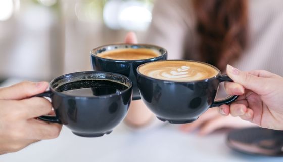 Drei Kaffeetassen in den Händen von Menschen. Im Hintergrund sind lange Haare und ein helles Oberteil zu erkenen.