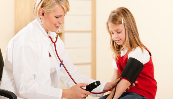 Ärztin misst Blutdruck bei einem Mädchen, das im roten T-Shirt neben ihr sitzt.
