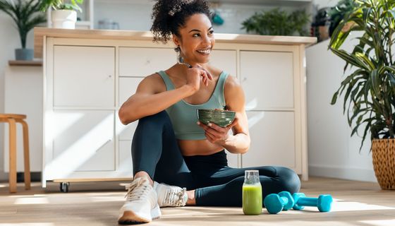 Junge Frau in Sportkleidung, neben sich Hanteln, sitzt auf dem Küchenboden, isst einen Salat.