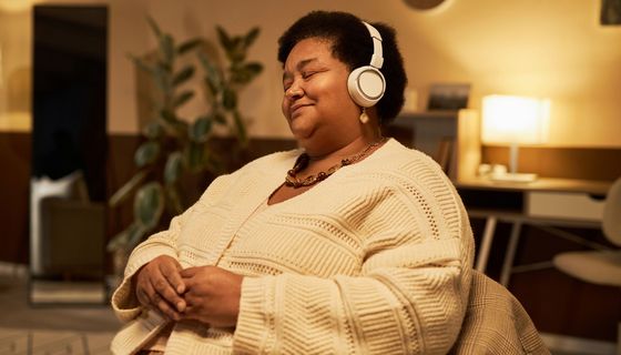 Eine Frau mittleren Alters sitzt auf einem Sessel. Sie lächelt und trägt Kopfhörer.