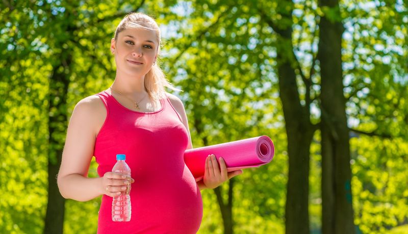 Eine schwangere Frau läuft mit Sportkleidung und Trinkflasche durch einen Park.