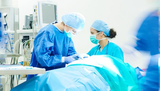 Patient liegt abgedeckt im Operationsaal, eine Chirurgin arbeitet am Patientin. Eine Op-Schwester überprüft die Werte.
