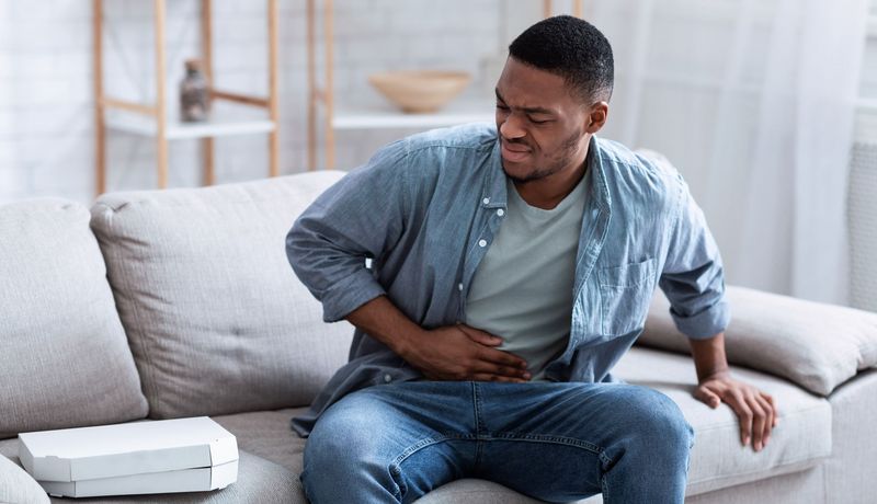 Ein junger Mann sitzt auf dem Sofa und hält sich den Bauch. Neben ihm liegt eine Pizza, die er leicht angeekelt betrachtet.