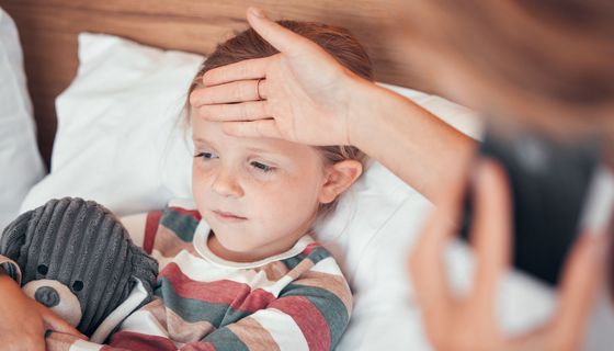 Ein kleines Mädchen liegt im Bett mit einem Kuscheltier in der Hand. Eine Person, die nicht erkennbar ist hält ihre Hand an die Stirn.