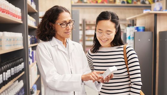 Eine Apothekerin hält ein Arzneimittel in der Hand und spricht mit einer zweiten Frau, die ebenfalls das Arzneimittel bertrachtet.