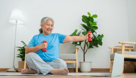 Eine ältere Frau sitzt in einem Wohnzimmer auf einer Sportmatte. Sie hat Hanteln in der Hand und schaut auf einen Laptop, der vor ihr steht.