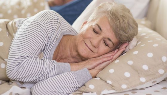 Eine ältere Dame schläft erholsam in einem Bett. Im Hintergrund ist ihr Ehemann zu sehen.