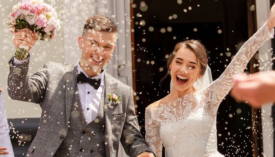 Hochzeitspaar, das gerade zur Kirchentür herauskommt: Der Bräutigam trägt den Brautstrauß, Gäste werfen Reis.