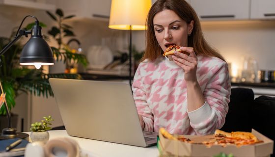 Frau sitzt abends vor dem Laptop und ist ein Stück Pizza.