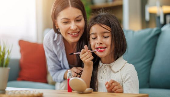 Ein Mädchen, etwa 10 Jahre, trägt Makeup auf und blickt in einen Handspiegel. Eine Frau sitzt neben ihr und schaut mit.