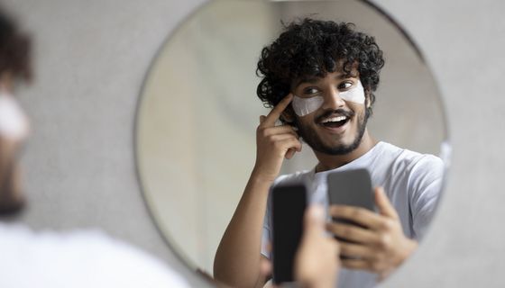 Junger Mann hat sein Handy in der Hand und blickt in den Spiegel. Auf seinen Wangen ist deutlich weiße Kosmetik zu sehen.