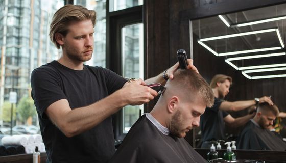 Ein Mann steht in einem Salon und rasiert einem anderen Mann den Kopf. Dieser hat den Kopf leicht nach vorne gebeugt.