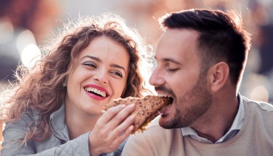 Junge Frau lässt einen Mann von ihrem Vollkorn-Brötchen abbeißen.