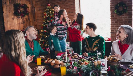 Acht Personen rund um den gedeckten Weihnachtstisch: Ein Kind sitzt auf den Schultern eines Mannes. Eine ältere Frau sitzt am Rand.