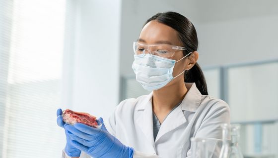 Eine junge Forscherin mit Maske und Handschuhen im weißen Kittel hält eine Petrischale mit Fleisch