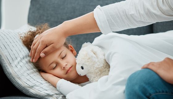 Krankes Kind liegt mit Kuscheltier auf dem Sofa und Mutter hält die Hand an die Stirn.