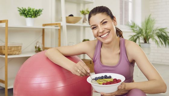Junge Frau mit Früchteteller in Sportkleidung, einen Arm auf einem Gymnastikball.