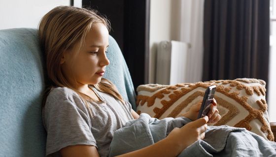 Ein junges Mädchen sitzt auf einem Sofa und hält lächelnd ein Smartphone in der Hand.
