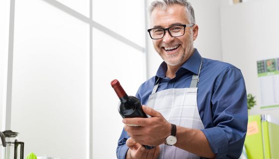 Älterer Mann in Schürze mit einer Weinflasche in der Hand, im Hintergrund eine Küche und Schneidebretter