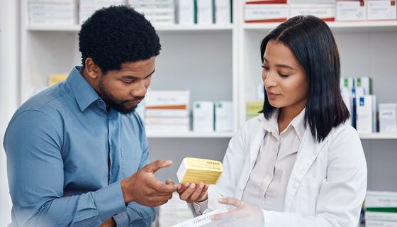 Ein dunkelhäutiger Mann steht neben einer Apothekerin im weißen Kittel und lässt sich beraten.