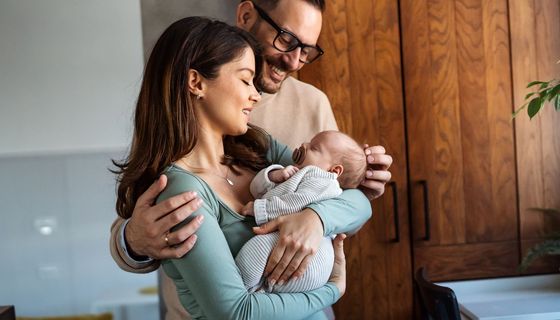 Eine Frau hält ein Baby, ein Mann hat seine Arme um sie gelegt und lächelt das Kind an.