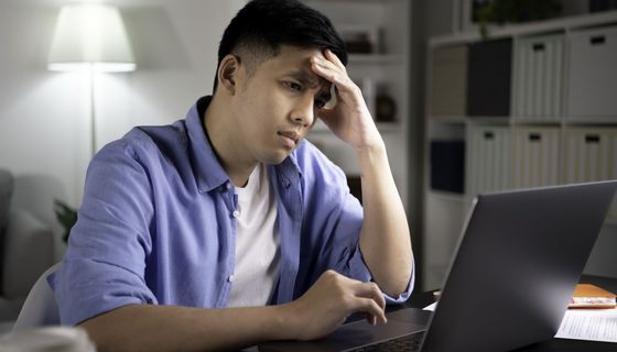 Junger Mann mit Augenringen hält sich die Stirn und blickt betroffen auf einen Laptopbildschirm.