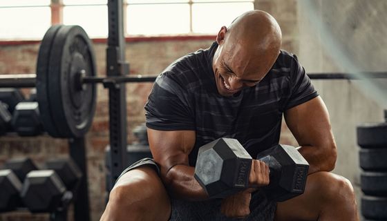 Ein Mann trainiert mit schmerzverrtem Gesicht in einem Fitnesstudio mit einer Hantel.