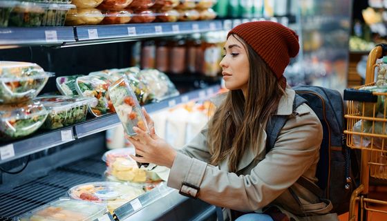 Junge Frau blickt prüfend auf einen Fertigsalat aus dem Kühlregal.