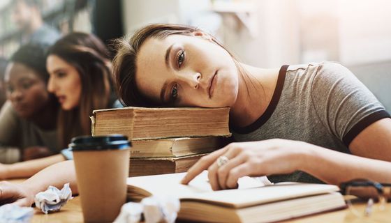 Traurige Studentin mit dem Kopf auf einem Stapel Bücher blickt glücklos in die Kamera. Im Hintergrund sind weitere Studenten verschwommen zu sehen.