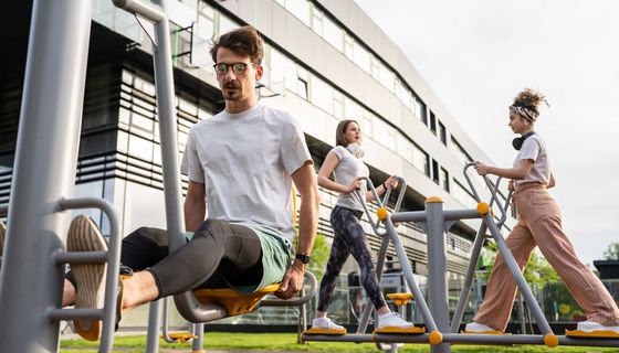 Ein Mann trainiert im Sitzen an einem Outdoor-Gerät, im Hintergrund sind zwei Frauen in Schrittstellung, ebenfalls auf Geräten, zu sehen.